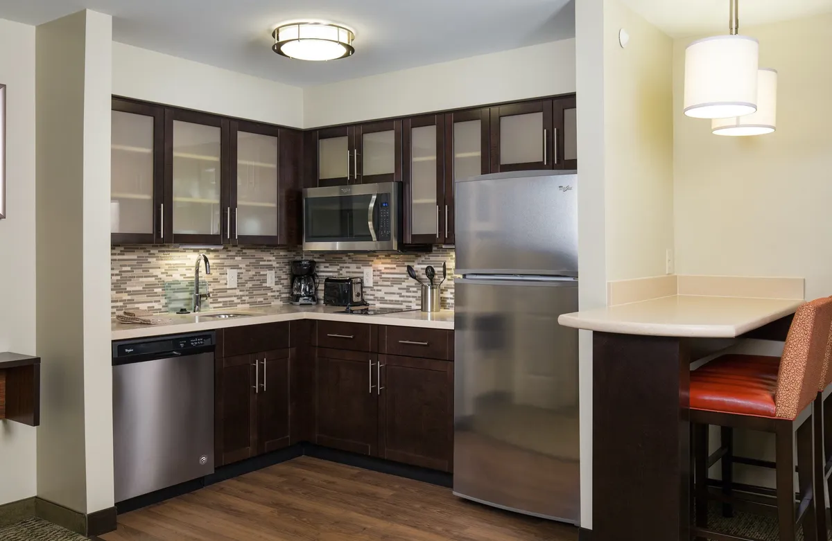 The image showcases a warm, inviting suite kitchen with modern stainless steel appliances, rich wooden cabinetry, and a sleek countertop bar that encourages guests to gather, cook, and socialize. The cozy ambiance is perfect for relaxing after a day exploring Denver.