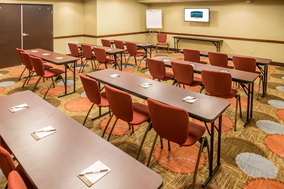 Inside, rows of tables are set up in a bright, contemporary space, focused on a presentation screen. The colorful carpet and layout suggest a productive environment for meetings, workshops, or training sessions.