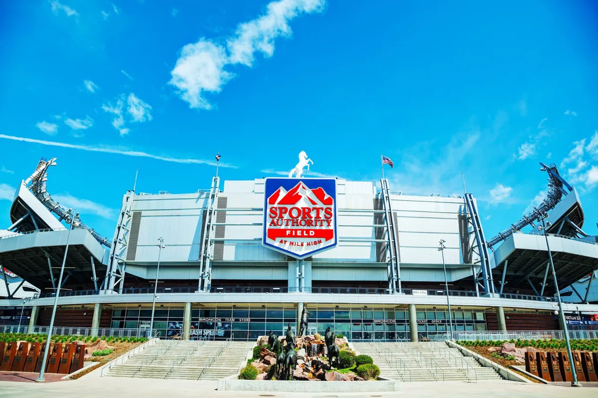 This image features the impressive exterior of a major Denver stadium, likely Empower Field at Mile High, with distinctive stadium signage, architectural details, and large sculptures in the foreground. The scene is lively and suggests a hub of sports events, concerts, and festive community gatherings, perfectly capturing the excitement and energy of Denver's city attractions.