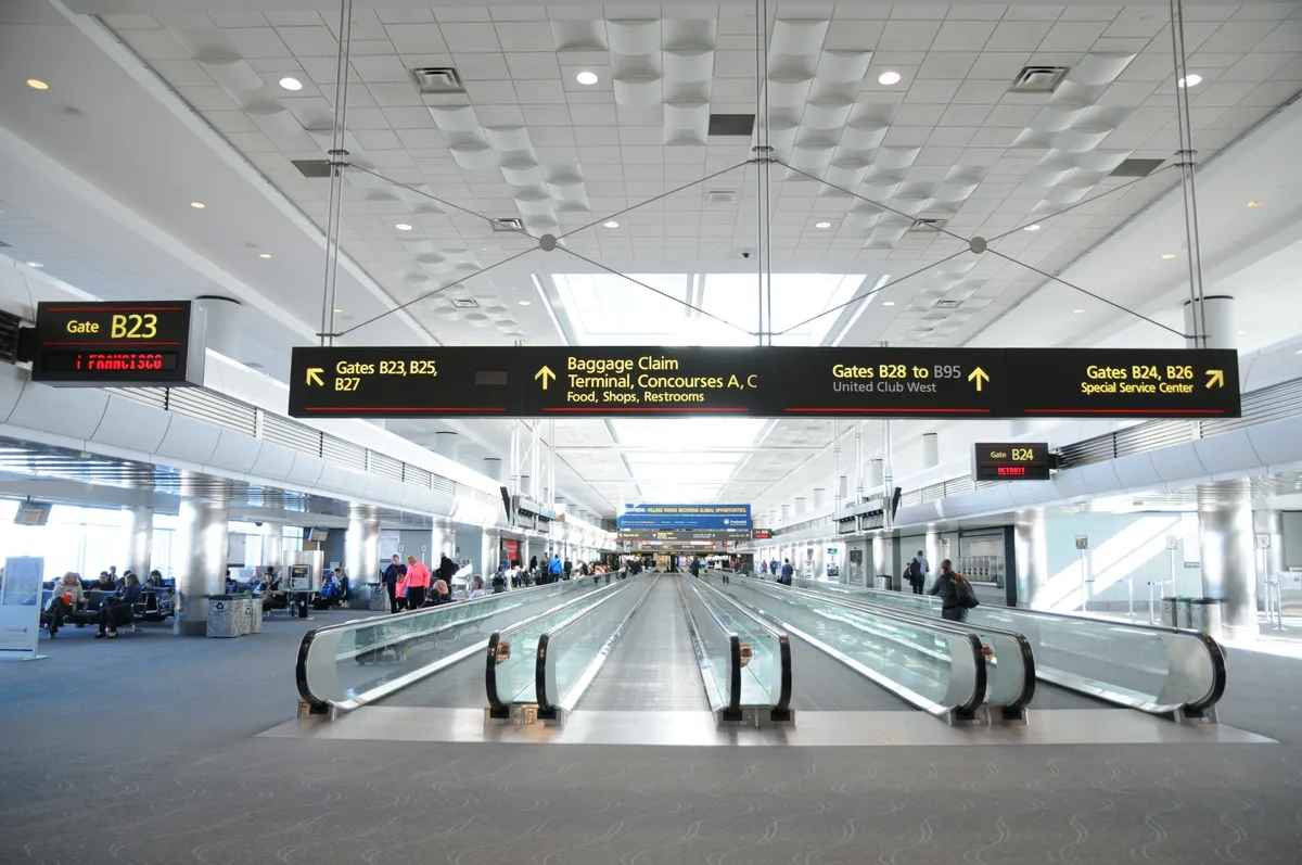 The image depicts a lively airport interior with moving walkways, information boards, and people waiting or moving about. There are families and groups gathered, some likely arriving or departing together, while others are shopping or dining at airport establishments. The overall ambiance is bustling and anticipatory, capturing the beginning of a trip or the excitement of reuniting with loved ones.