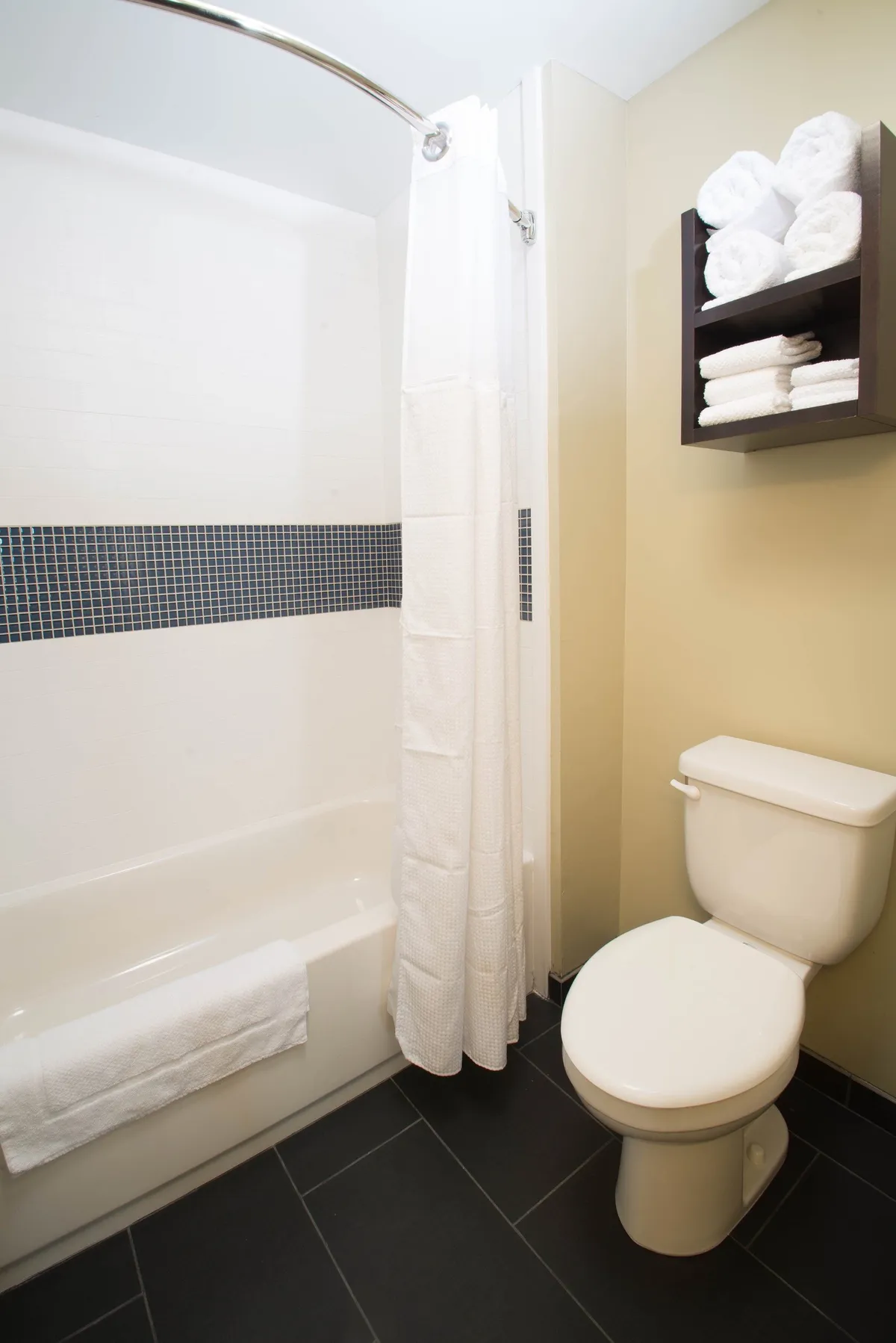 A calming, well-lit bathroom featuring a bathtub with a shower curtain, a towel rack stacked with fluffy rolled towels, and a toilet. The space exudes cleanliness and comfort, perfect for relaxing self-care routines.