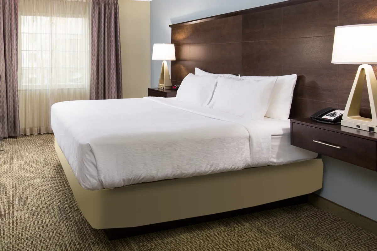 A tranquil king-sized bed is centered in the suite's bedroom, flanked by a nightstand with a lamp. Soft, sheer curtains allow natural morning light to filter in, creating a peaceful and restful ambiance ideal for relaxation.