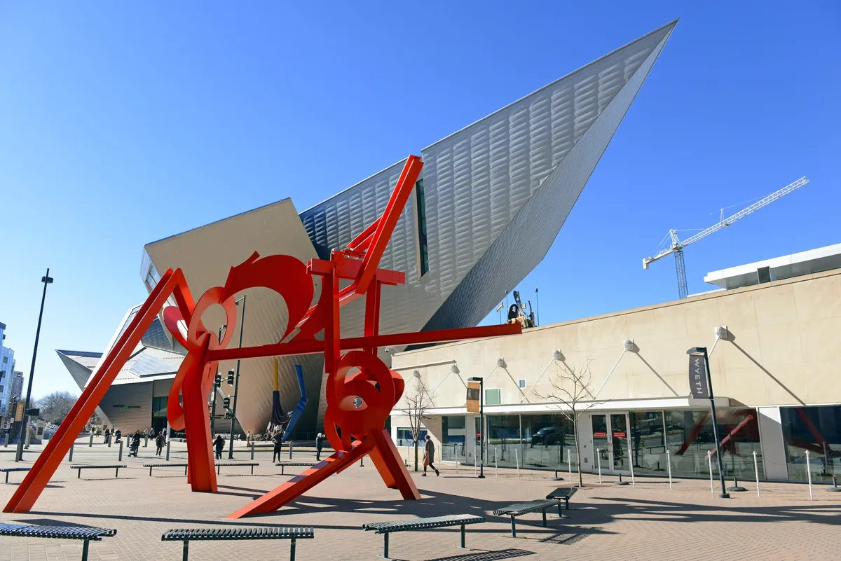 The scene captures a modern architectural building with a striking large red sculpture under a clear blue sky. The public space is open and inviting, evoking creativity and dynamic urban energy.